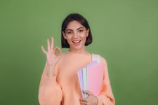 Smiliing young girl student doing an okay sign holding her books in her arm