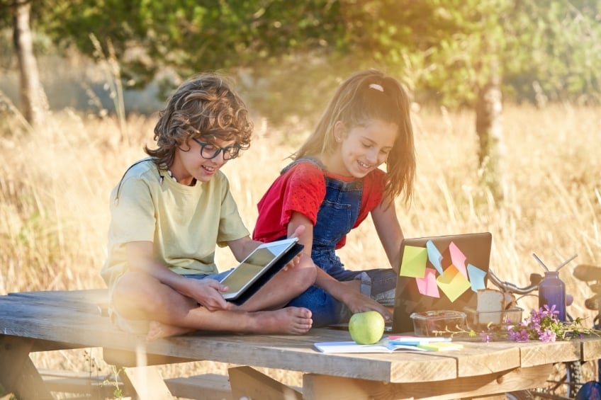 2 children sitting on table outdoors in summer using their tablets to prevent the summer slide